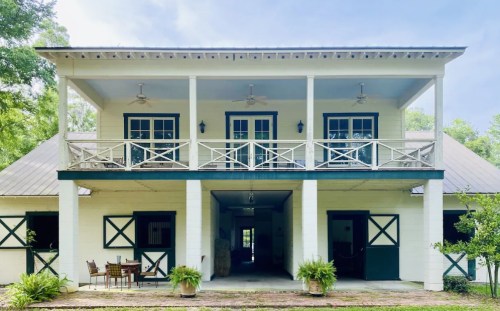 A two-story house with a balcony, white walls, and green accents, surrounded by greenery and featuring outdoor seating.