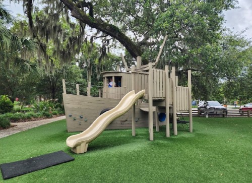 A wooden playground structure resembling a ship, featuring a slide and climbing elements, surrounded by greenery.