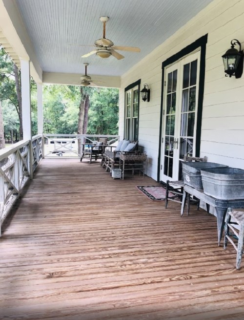 A spacious porch with wooden flooring, rocking chairs, and a vintage wash basin, surrounded by greenery.
