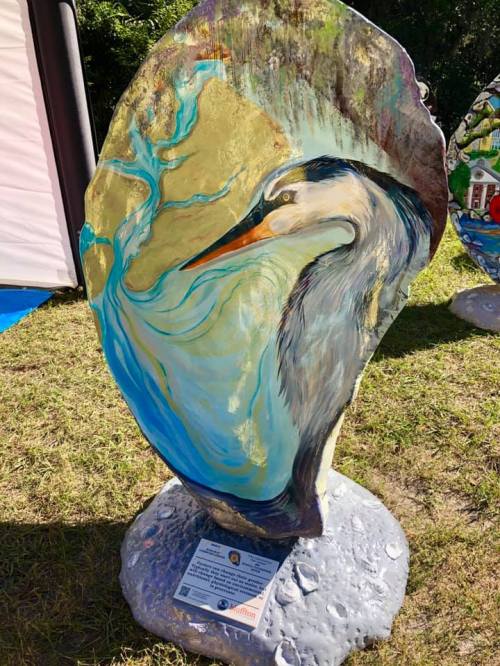 A painted stone sculpture of a heron, featuring vibrant colors and flowing designs, displayed outdoors on grass.