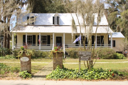 A charming two-story house with a porch, surrounded by greenery, featuring a sign that reads "Welcome Center."