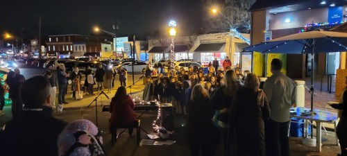 A crowd gathers at night for an outdoor event, with a performer at a table and festive lights in the background.