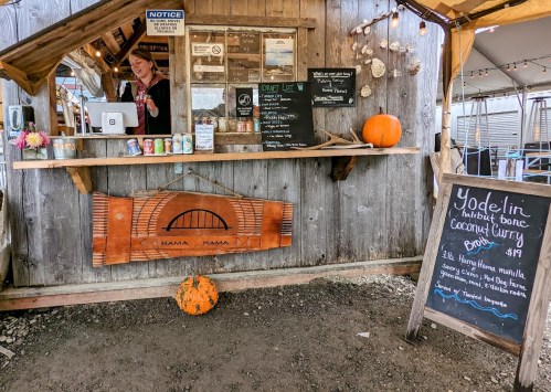 A rustic food stall with a menu board, a smiling worker, and a pumpkin on display. Cozy outdoor setting.