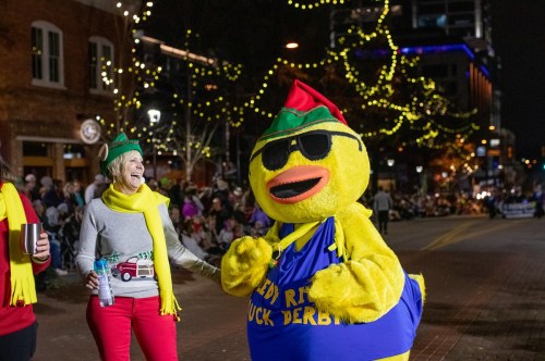 A person in a festive outfit laughs with a large, cheerful duck mascot wearing sunglasses and a holiday-themed costume.
