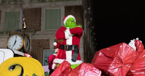 The Grinch in a Santa suit stands on a float surrounded by large red bags, with a clock tower in the background.