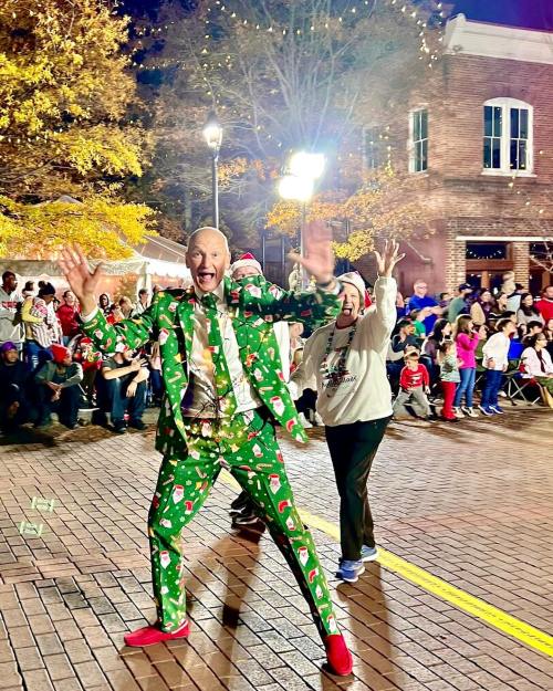 A man in a festive green suit with holiday patterns joyfully waves, while a woman in a sweater smiles behind him at a parade.