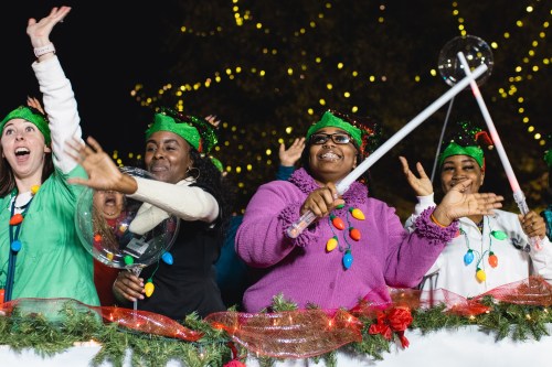 A group of joyful women in festive attire celebrate with smiles and props during a holiday event, surrounded by lights.