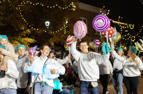A festive parade scene with people in matching sweatshirts, holding colorful lollipops and smiling under string lights.