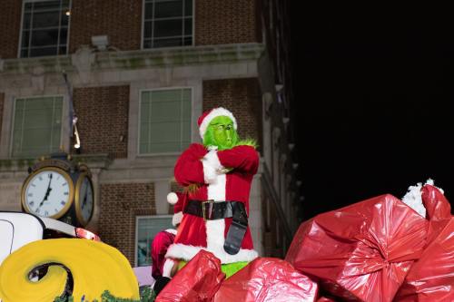 The Grinch in a Santa suit stands on a festive float, surrounded by large red gifts, against a night sky.