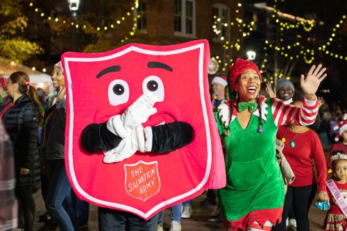 A festive scene with a large Salvation Army mascot and a woman in a holiday outfit, both joyfully celebrating in a crowd.