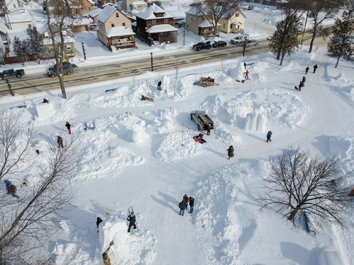 Aerial view of a snowy park with people building snow sculptures and playing in the snow, surrounded by houses and trees.