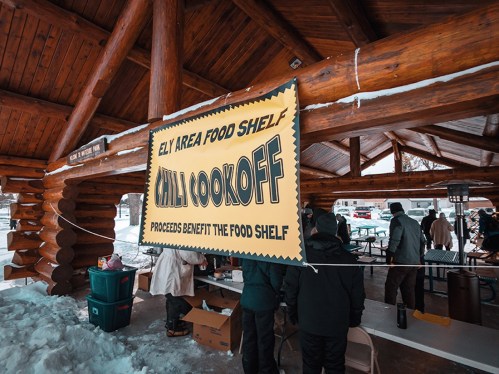 A banner for a chili cook-off hangs in a snowy outdoor pavilion, promoting benefits for the Ely Area Food Shelf.