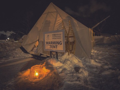 A warming tent with a sign inviting people inside, surrounded by snow and a glowing lantern at night.