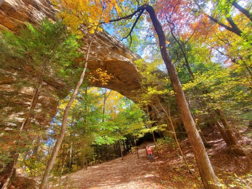 A natural stone arch surrounded by vibrant autumn foliage and a person walking along a forest path.