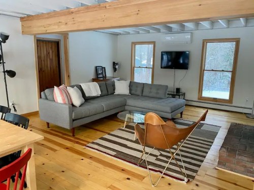 Cozy living room with a gray sectional sofa, wooden floors, and large windows, featuring a TV and stylish decor.