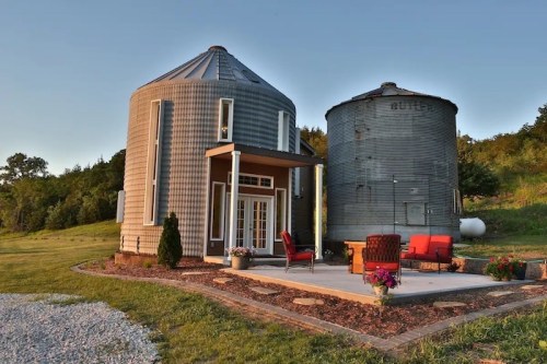 Two converted silos serve as a unique home, featuring a patio with seating and surrounded by greenery.