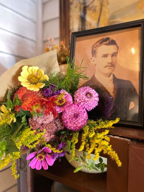 A vintage photo of a man beside a colorful bouquet of flowers in a vase on a wooden shelf.