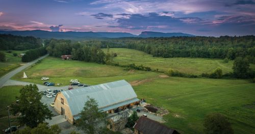 A scenic view of a barn surrounded by green fields and mountains under a colorful sunset sky.