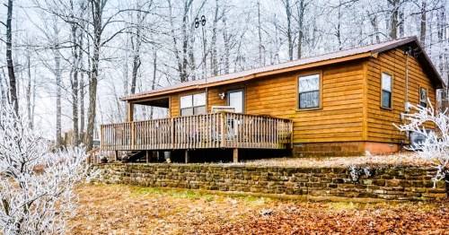 A cozy wooden cabin surrounded by frosty trees and a stone wall, with a deck in front and autumn leaves scattered around.