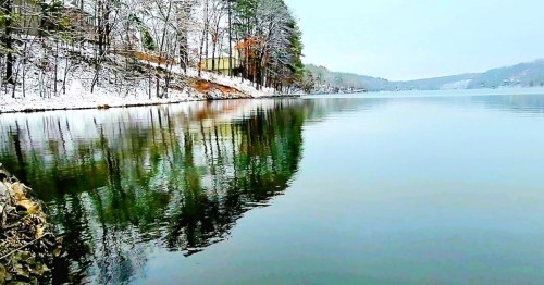 A serene winter landscape with snow-covered trees reflecting on calm water.