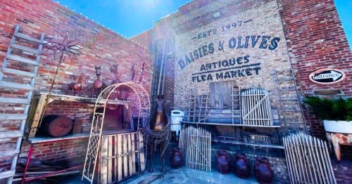 A rustic flea market entrance with vintage items and a brick wall featuring "Daisies & Olives Antiques."