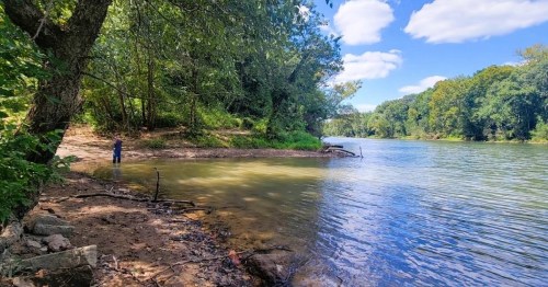 A serene river scene with a person standing by the water's edge, surrounded by lush greenery and a clear blue sky.