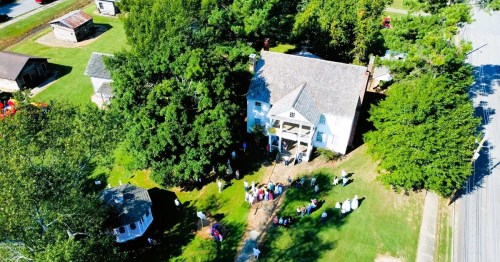 Aerial view of a historic house surrounded by trees, with a gathering of people on the lawn and small buildings nearby.