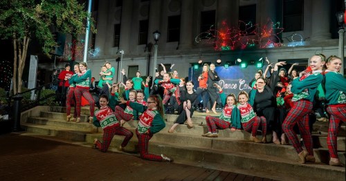 A lively dance group in festive outfits poses on steps, with holiday lights and decorations in the background.