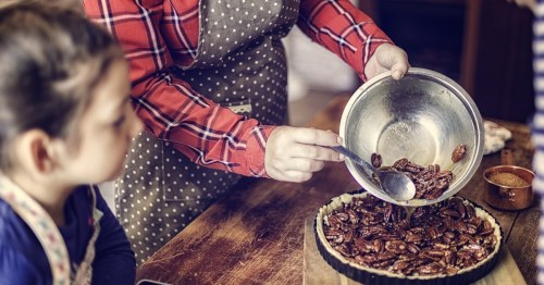 A person in an apron adds pecans to a pie crust while a child watches attentively.