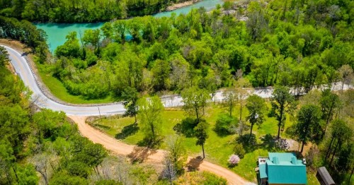 Aerial view of a winding road through lush green trees near a river, with a house visible in the foreground.