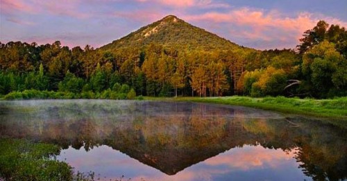 A serene landscape featuring a mountain reflected in a calm lake, surrounded by lush trees and a colorful sky at sunset.