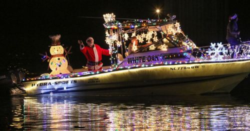 A festive boat decorated with lights, featuring Santa waving and a snowman on display, gliding on water at night.