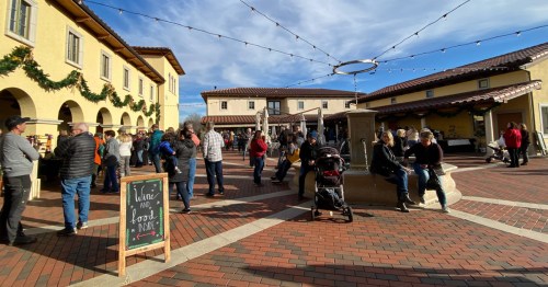 A bustling outdoor market scene with people gathered around a fountain, string lights overhead, and festive decorations.