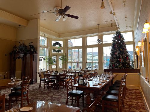 Cozy restaurant interior with wooden tables, chairs, and a decorated Christmas tree by large windows.