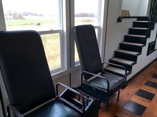 Two black chairs by large windows, with a staircase nearby and a view of a field and horse outside.