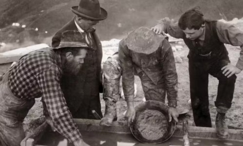 Four men panning for gold in a historical setting, focused on the pan and surrounded by mining equipment.