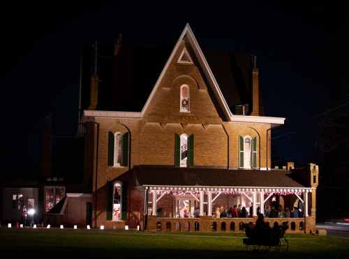 A beautifully lit historic house at night, featuring a porch and festive decorations, with people gathered outside.