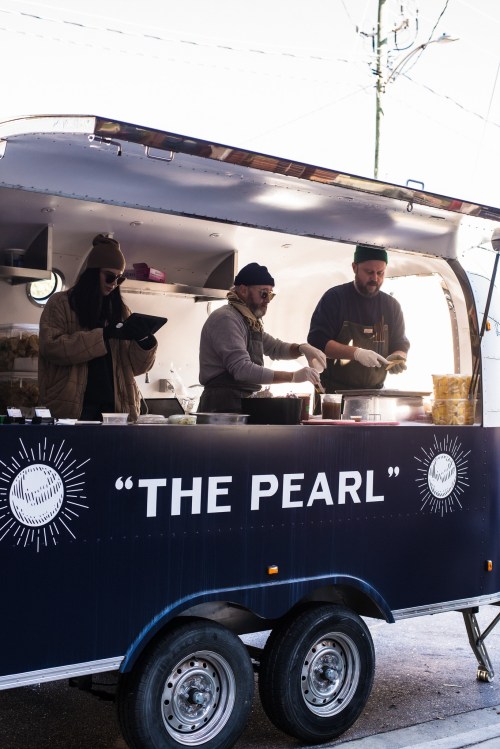 A food truck named "The Pearl" with three people preparing food inside. Bright sunlight illuminates the scene.