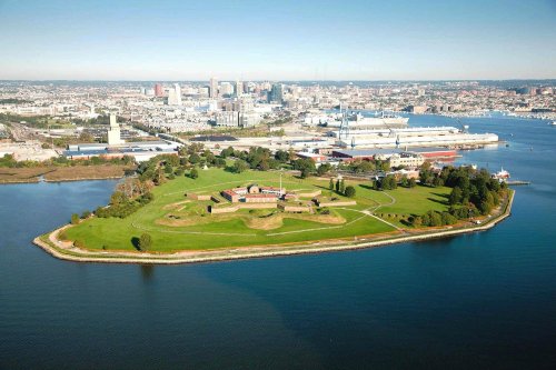 Aerial view of a green island with historic buildings, surrounded by water and a city skyline in the background.