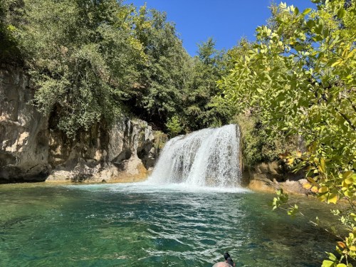 A serene waterfall cascades into a clear blue pool, surrounded by lush greenery and a bright blue sky.