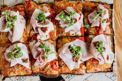 A close-up of a rectangular pizza topped with slices of ham, herbs, and roasted red peppers on a wooden surface.