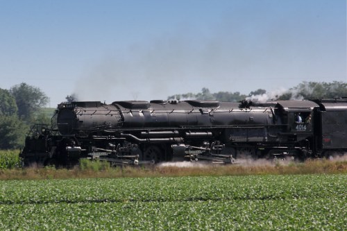 A vintage steam locomotive chugs through a green field, releasing steam into the clear blue sky.