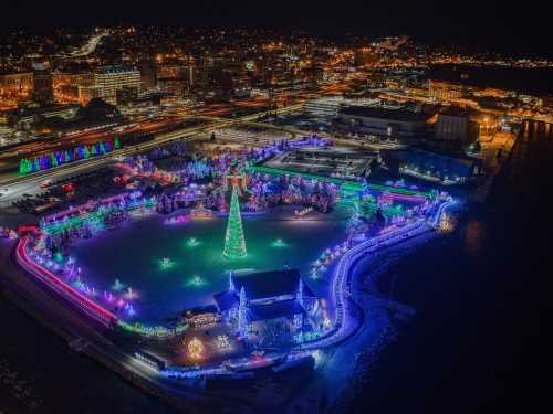 Aerial view of a festive park illuminated with colorful Christmas lights, featuring a large tree and snowy landscape at night.