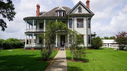 A large, historic Victorian-style house with a green lawn and archway of plants in the foreground.