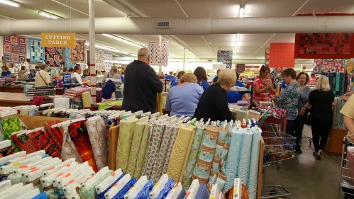 A busy fabric store with shoppers browsing colorful fabrics and a cutting table in the background.