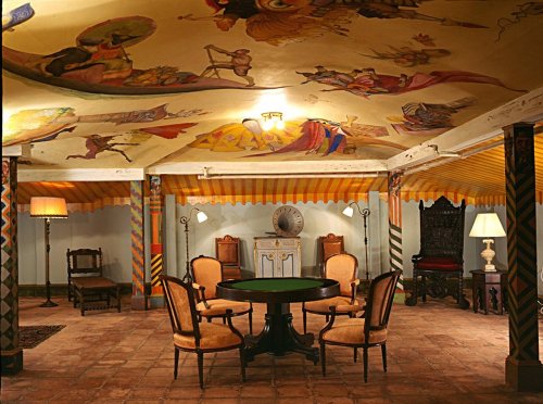 A vintage room with a painted ceiling, featuring a round table and elegant chairs, illuminated by lamps.
