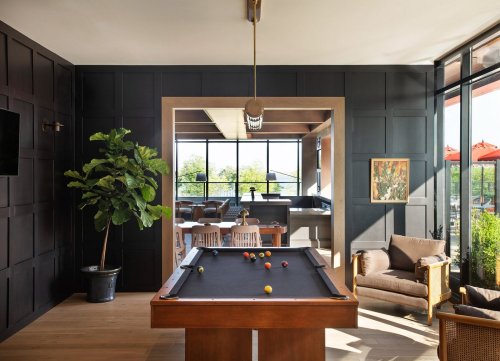 A modern game room featuring a pool table, large windows, and stylish seating with greenery in the corner.