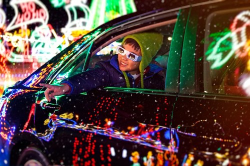 A joyful child in a car points out colorful holiday lights, wearing 3D glasses and a bright green beanie.