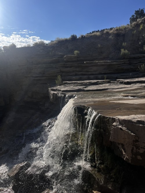 A waterfall cascades over rocky ledges under a bright blue sky with scattered clouds.
