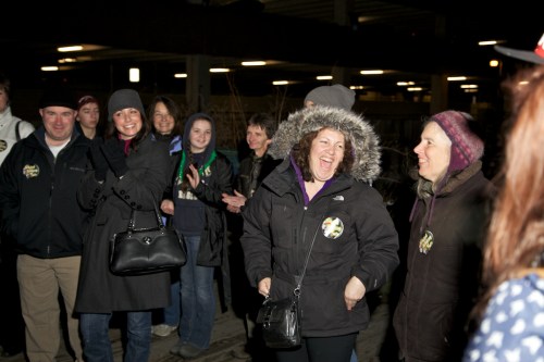 A group of people laughing and chatting together in a dimly lit indoor space, wearing winter clothing.